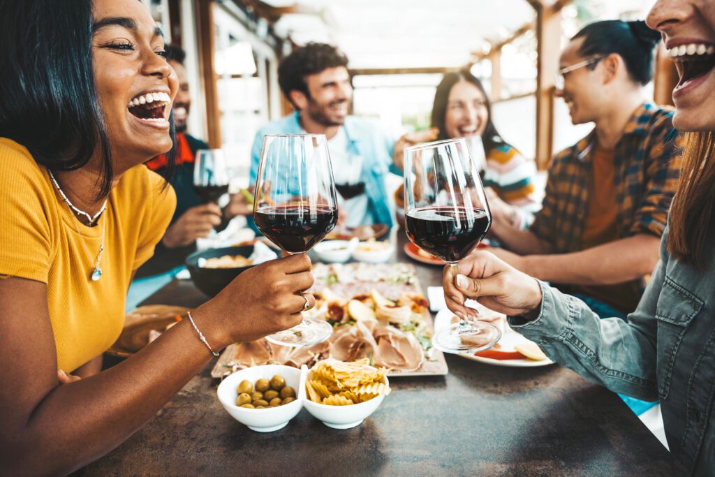 Happy friends drinking red wine sitting at restaurant table Multiracial young people enjoying rooftop dinner party together Food and beverage concept with guys and girls having lunch break outside