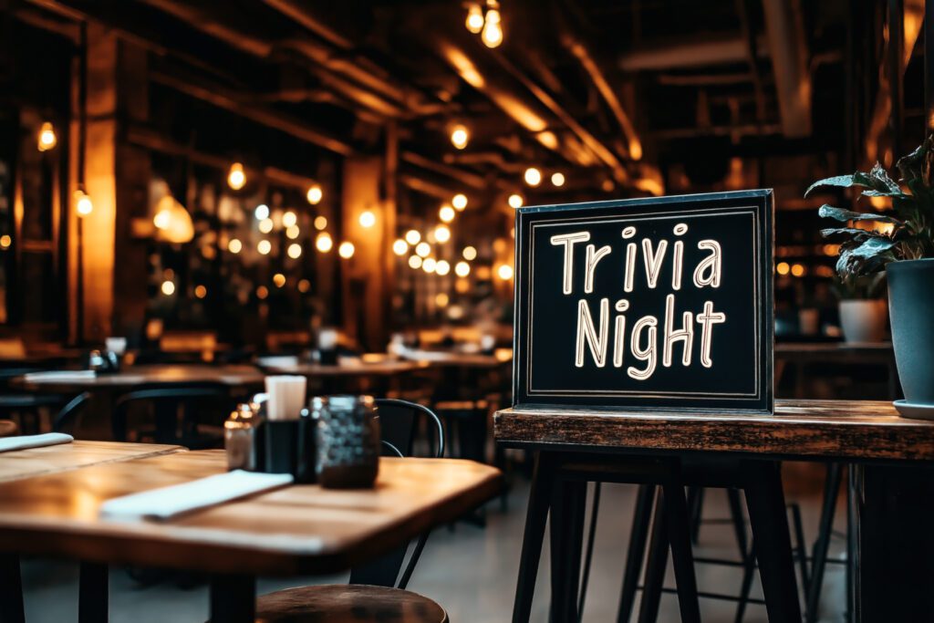 Cozy, dimly lit pub or restaurant interior with a sign on a table that reads 'Trivia Night'. The background includes tables, chairs, and warm decorative lighting.